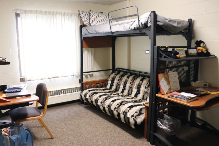 A dorm room with a lofted bed above a futon. The bed has a gray comforter and patterned pillows, while the futon has a black and beige patterned cover. To the right, there is a desk with books, papers, and framed photos on the top shelf. A wooden desk and chair are on the left side.